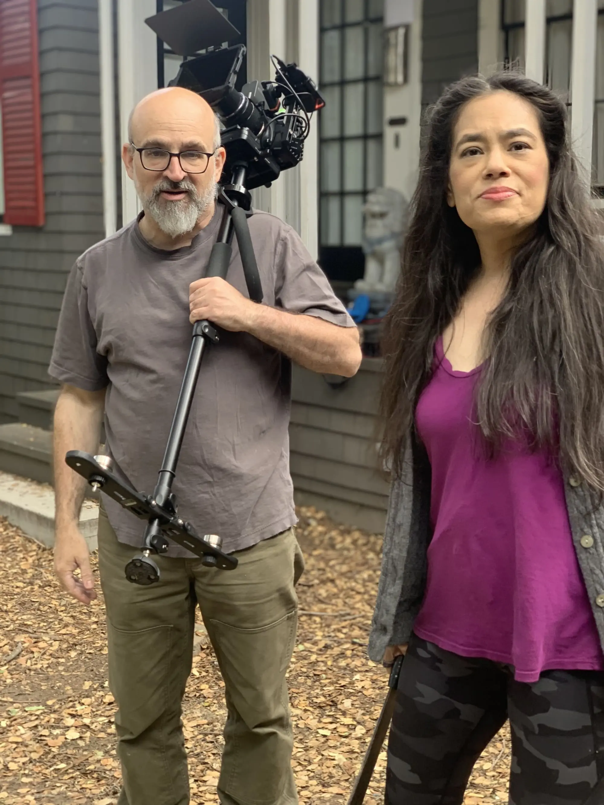 Outdoors by a building with dark siding and red shutters, a man with a camera stabilizer stands beside a woman for Old Bat Films.