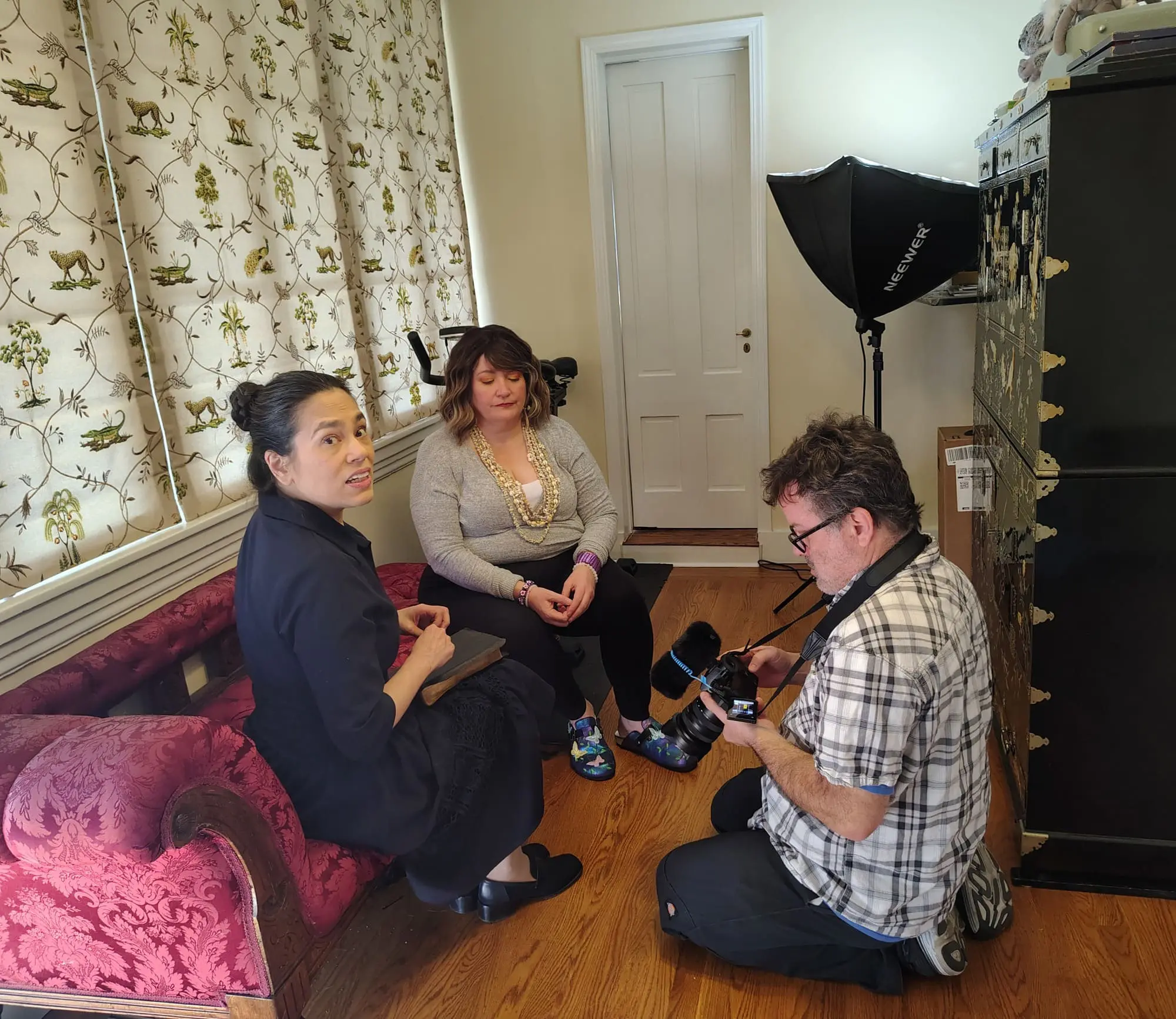 On a red couch, two women sit as a man kneels adjusting a camera; Old Bat Films equipment and ornate cabinet are in the room.