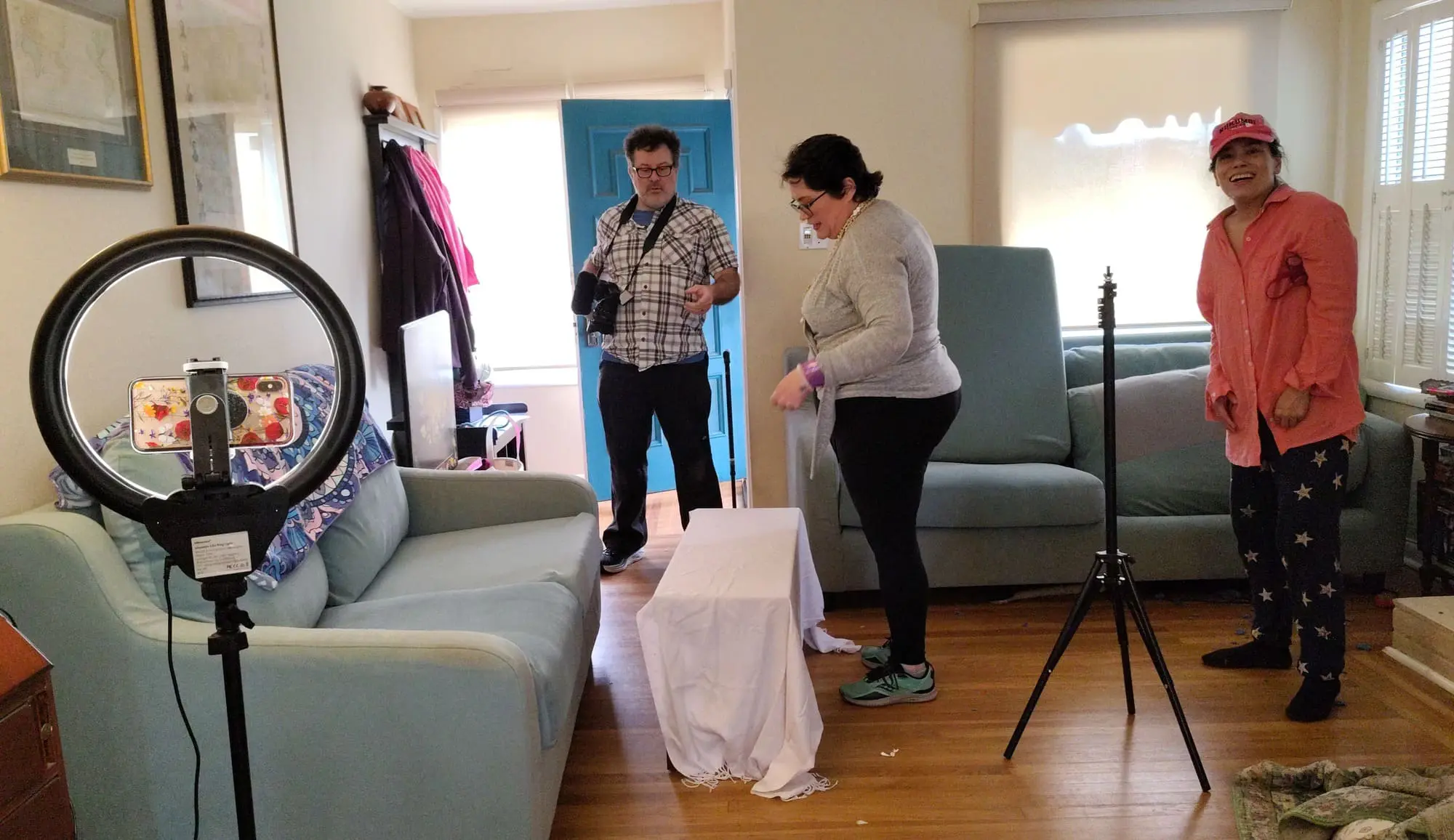 In a living room, three people set up a ring light, phone on tripod, and white-clothed table for Old Bat Films.