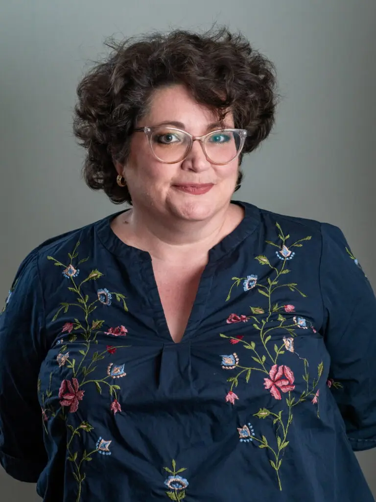 Elizabeth Butler of Old Bat Films stands against a gray background, wearing glasses and a blue floral blouse, slightly smiling.