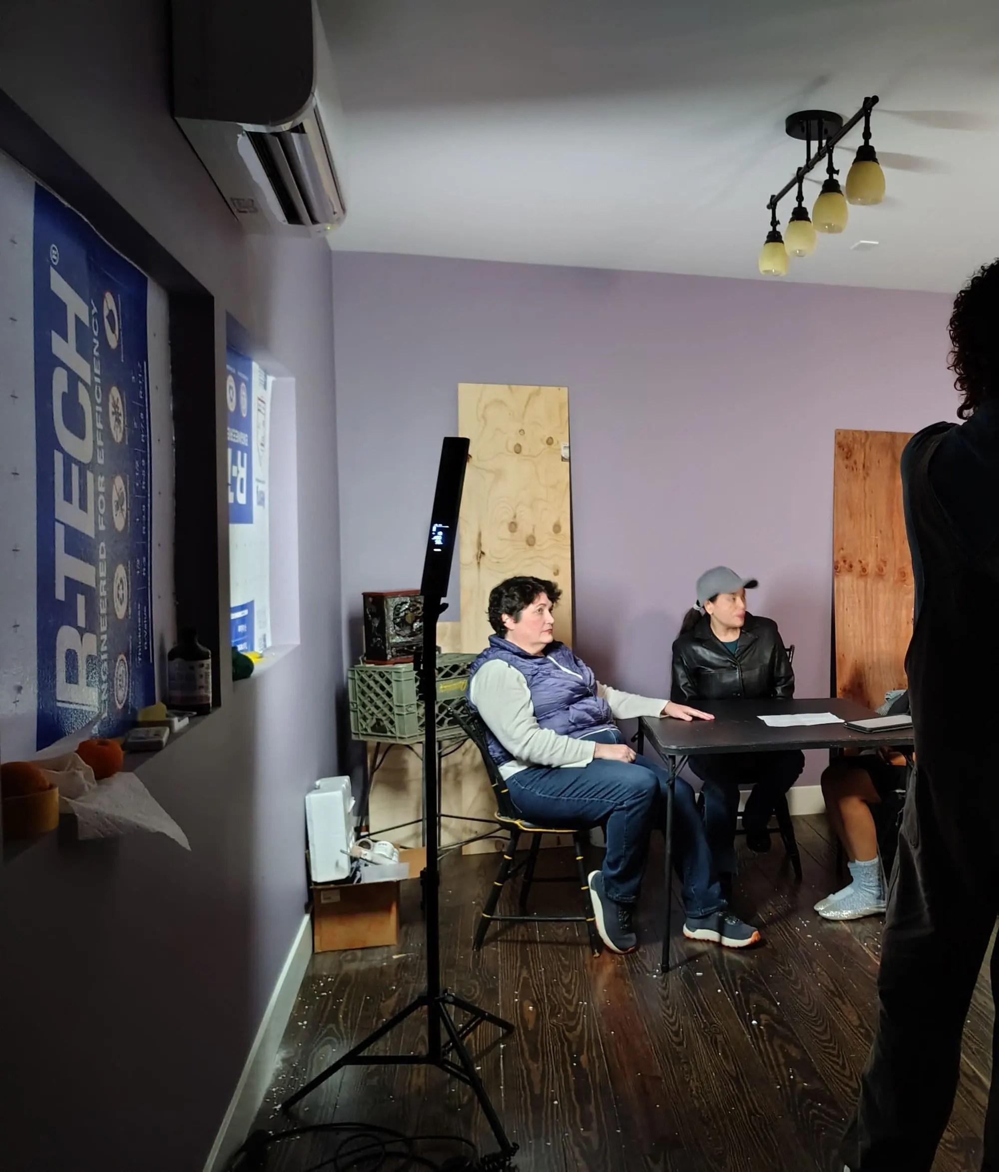 In an Old Bat Films scene, two people sit at a table in a purple-walled room as another stands nearby amid construction materials.