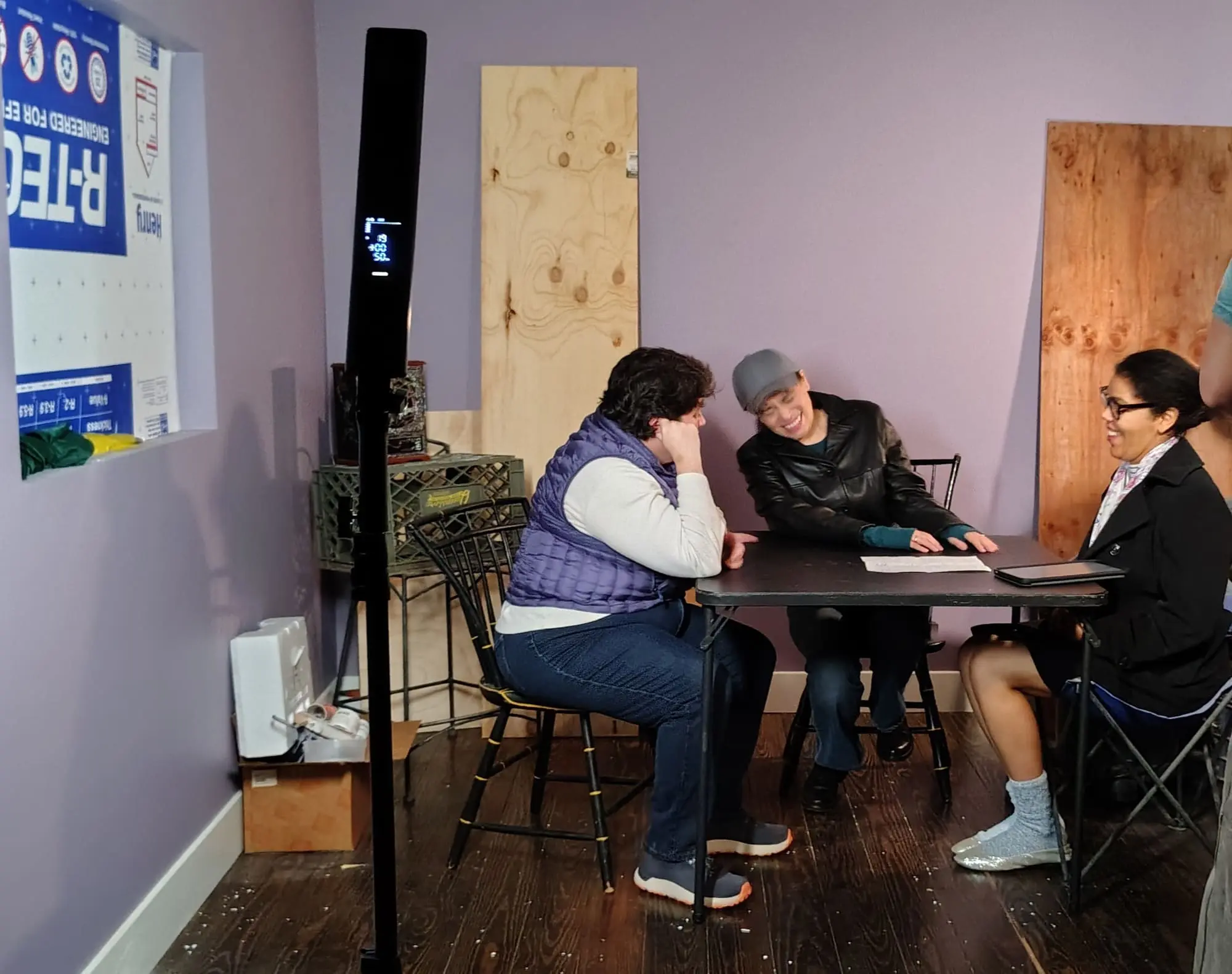 In a small purple-walled room, three people talk at a black table near a tall object; Old Bat Films decor accents the scene.
